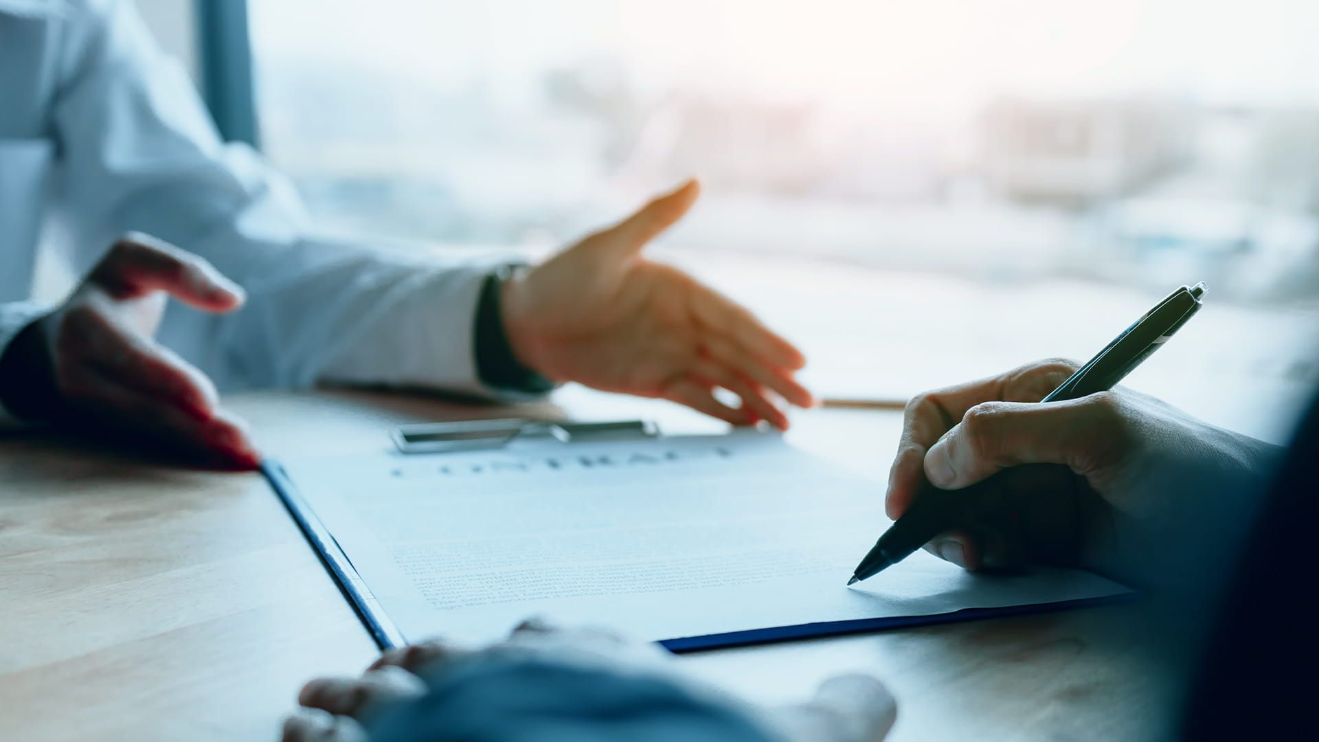 Close-up of a person signing a document on a clipboard while another hand gestures across the table.