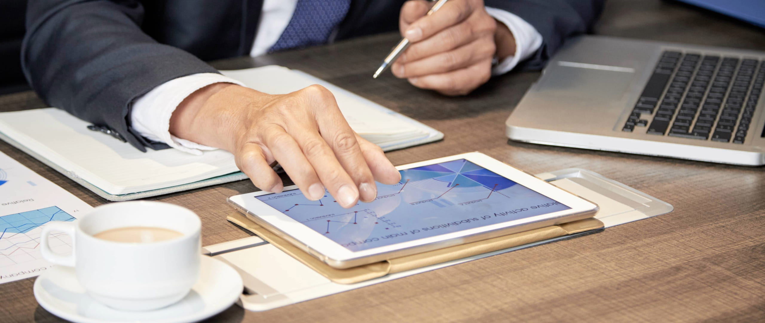 Business desk scene with tablet displaying charts, laptop, documents, and coffee cup.